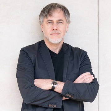 Man with short gray hair and beard wearing a dark blazer, standing with arms crossed against a light wall.