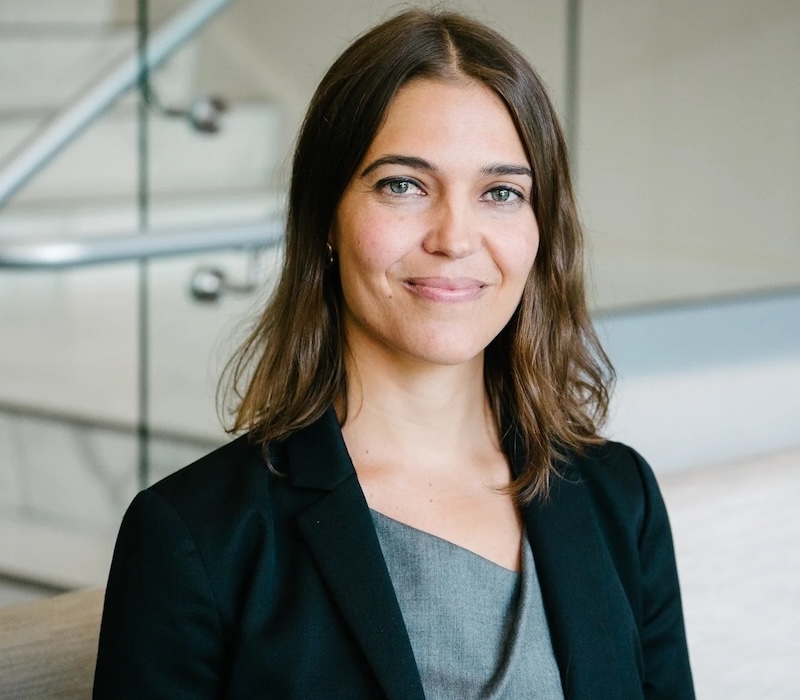 Smiling woman with shoulder-length brown hair wearing a gray top and black blazer, seated in a modern indoor setting with glass railings in the background.