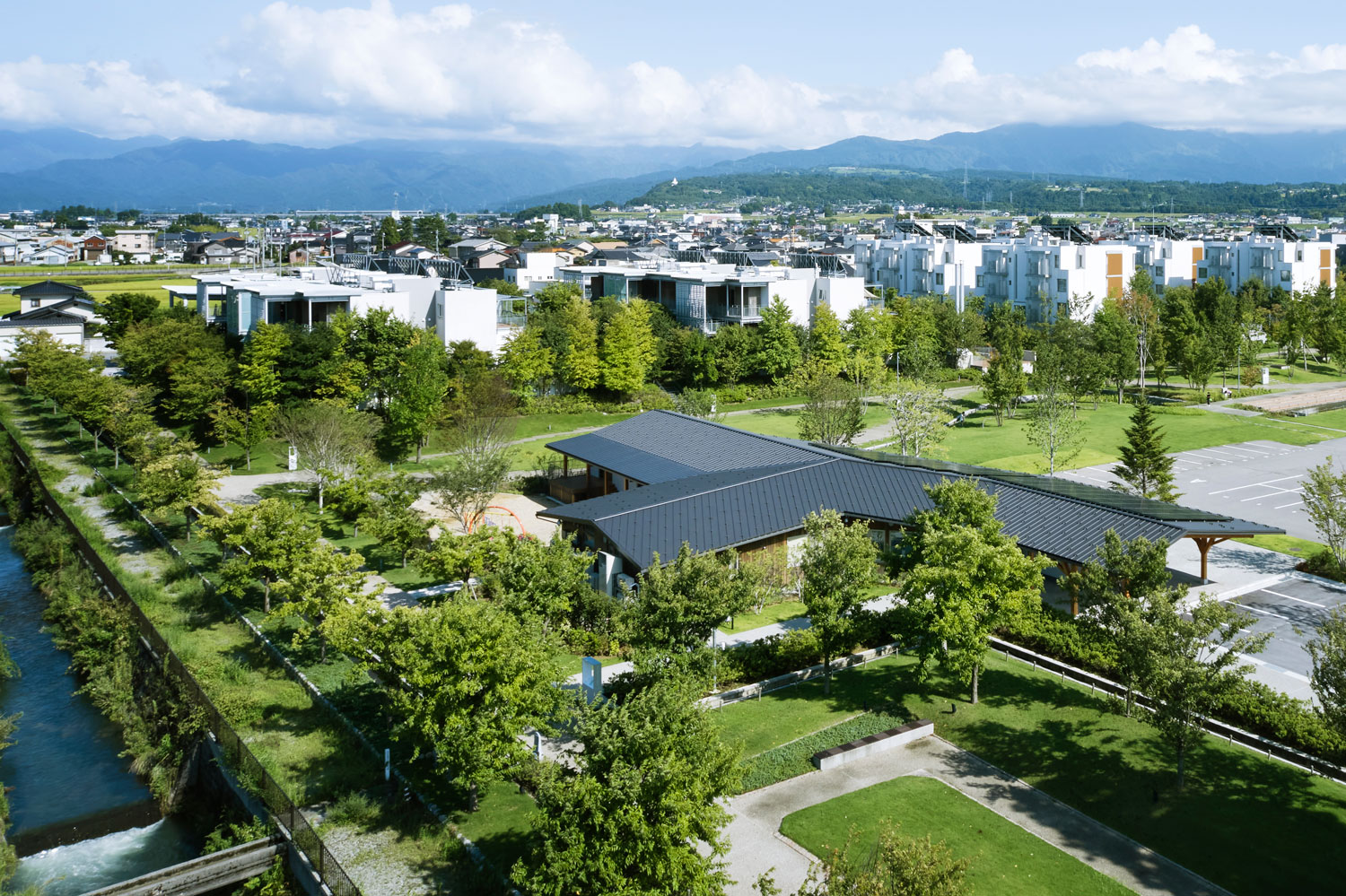 A pavilion in a green park next to a waterway. In the background is a low-rise housing complex. Passive Town by Placemedia.