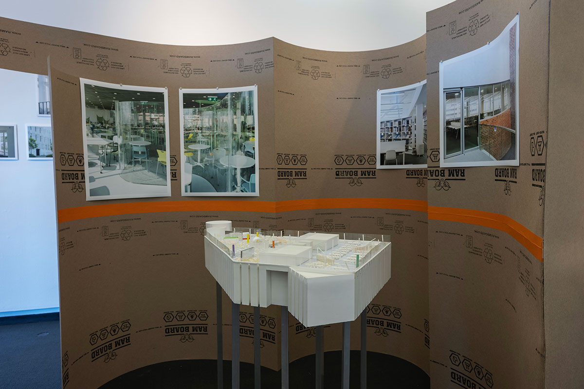 A curved brown wall displays multiple images, with a freestanding architectural model in front.