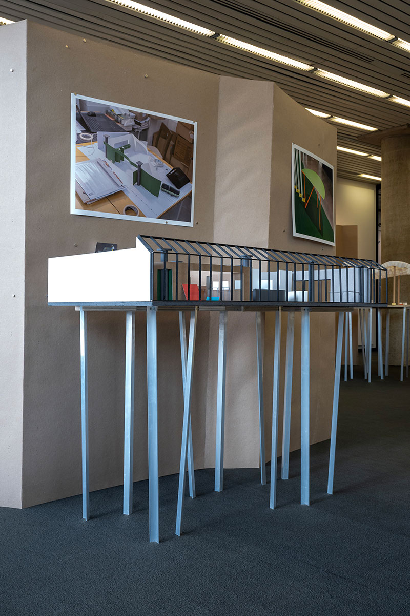 An architectural model of a building displayed on aluminum legs in front of a temporary wall with images.