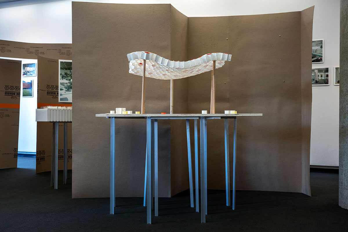 An architectural model of a curved lattice shape displayed on aluminum legs in front of a curved temporary wall made of chipboard.