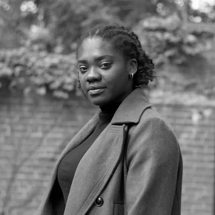 Black-and-white portrait of a woman with short curly hair wearing a coat and turtleneck, standing outdoors in front of a brick wall.