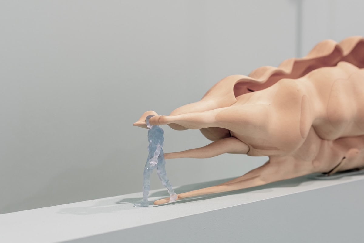 Abstract sculpture of a fleshy, organic form resting on a ledge, with a small translucent blue figure emerging and walking from one of its fingers.