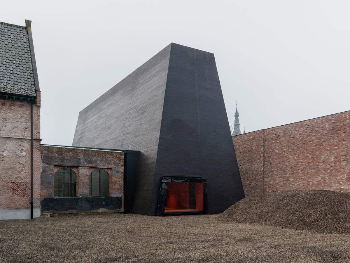 Angular dark-brick modern building with a red-lit entrance, set between older brick structures in a gravel courtyard under an overcast sky.