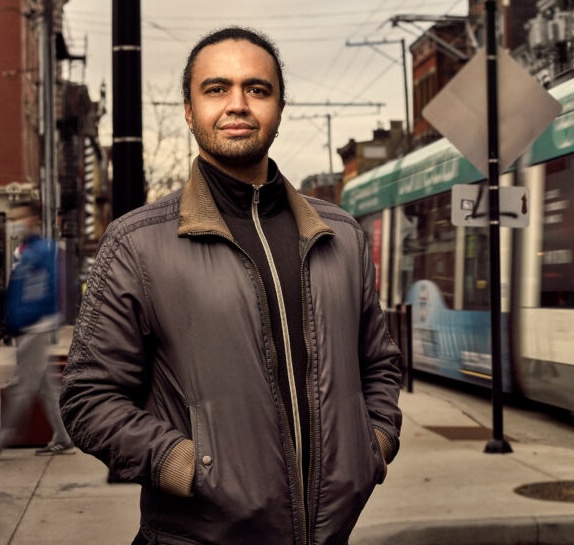 Person in a gray jacket stands on a city sidewalk with a streetcar passing in the background.