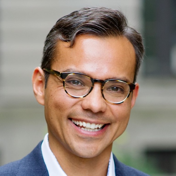 Smiling man with short dark hair and glasses wearing a suit jacket, photographed outdoors with a blurred background.