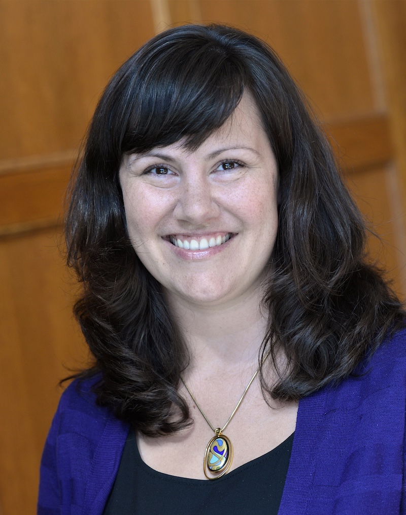 Smiling woman with long dark wavy hair wearing a purple cardigan, black top, and colorful pendant necklace against a wooden background.