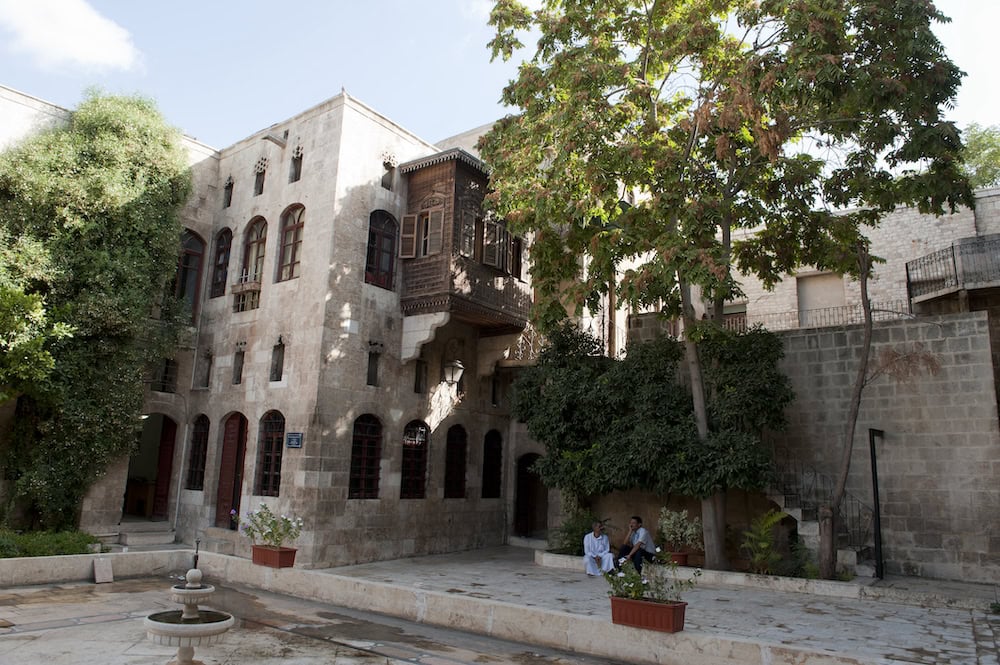 Exterior of old city building in Aleppo, Syria with two men sitting outside. "Aleppo Old City Rehabilitation Office 0101" by Dosseman is licensed under CC BY-SA 4.0.