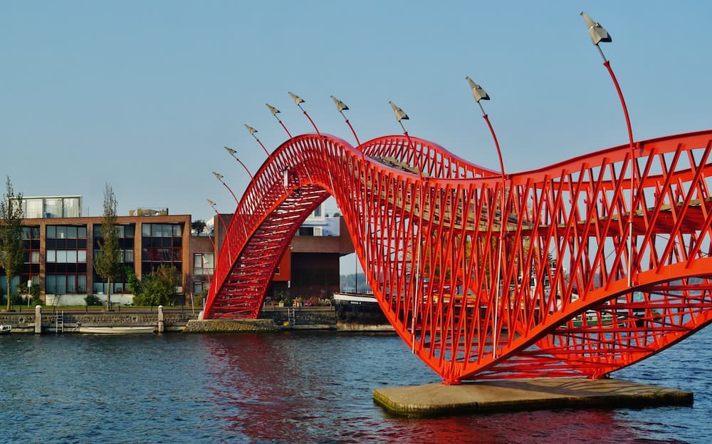 Photo of a red metal bridge extending over a river in Amsterdam. "Amsterdam Python Bridge 14" by Zairon is licensed under CC BY-SA 4.0.