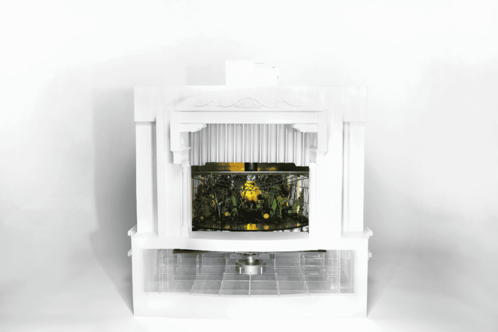 A model of a white opera set, with a motor underneath it and a lush green scene onstage.