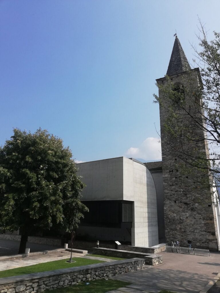 Exterior of parish complex and Church of Monte Carasso. "File:Convento delle agostiniane a Monte Carasso, progetto di Luigi Snozzi, veduta con campanile della chiesa dei santi Bernardino e Girolamo.jpg" by Flautor is marked with CC0 1.0.