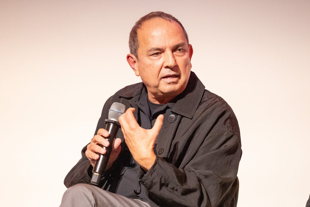 Man with dark shirt talks into microphone while seated.