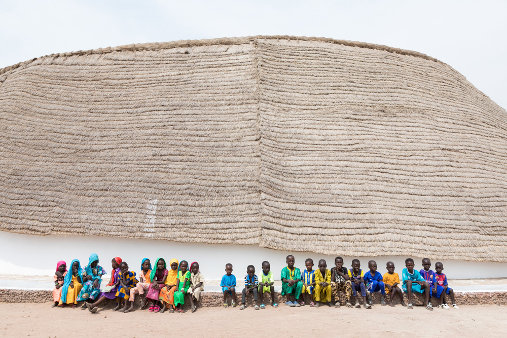 A group of children sit beside a structure with a large roof made of organic materials.