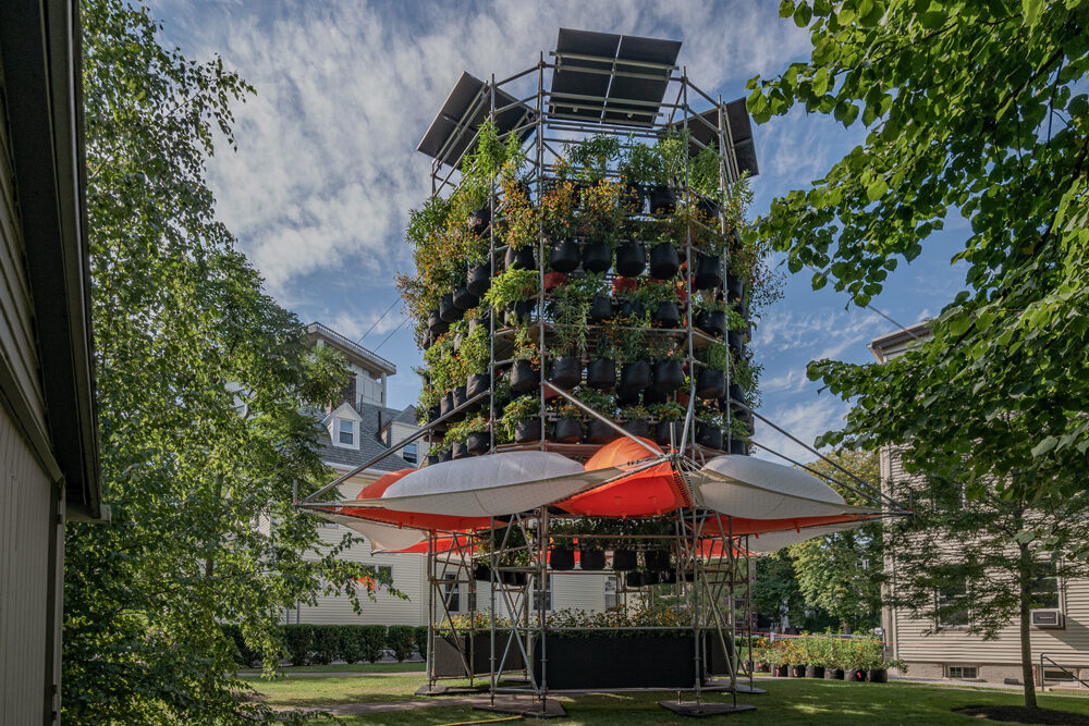 A photograph of Belinda Tato's Polinature project. A four-story cylindrical structure of scaffolding supports tiers of plants in black grow bags. Orange and white inflated bubbles ring the lower level.