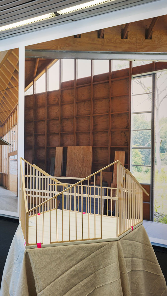 A wooden architectural model on a pedestal draped with canvas, in front of a wall with large images of wooden interior walls.