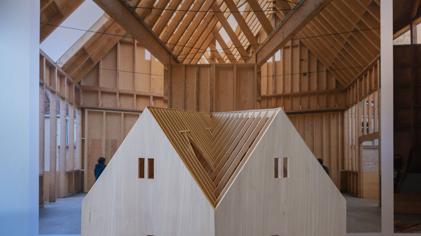 Detail of a wooden architectural model with an angled roof in front of a large mural image of wooden interior framing.