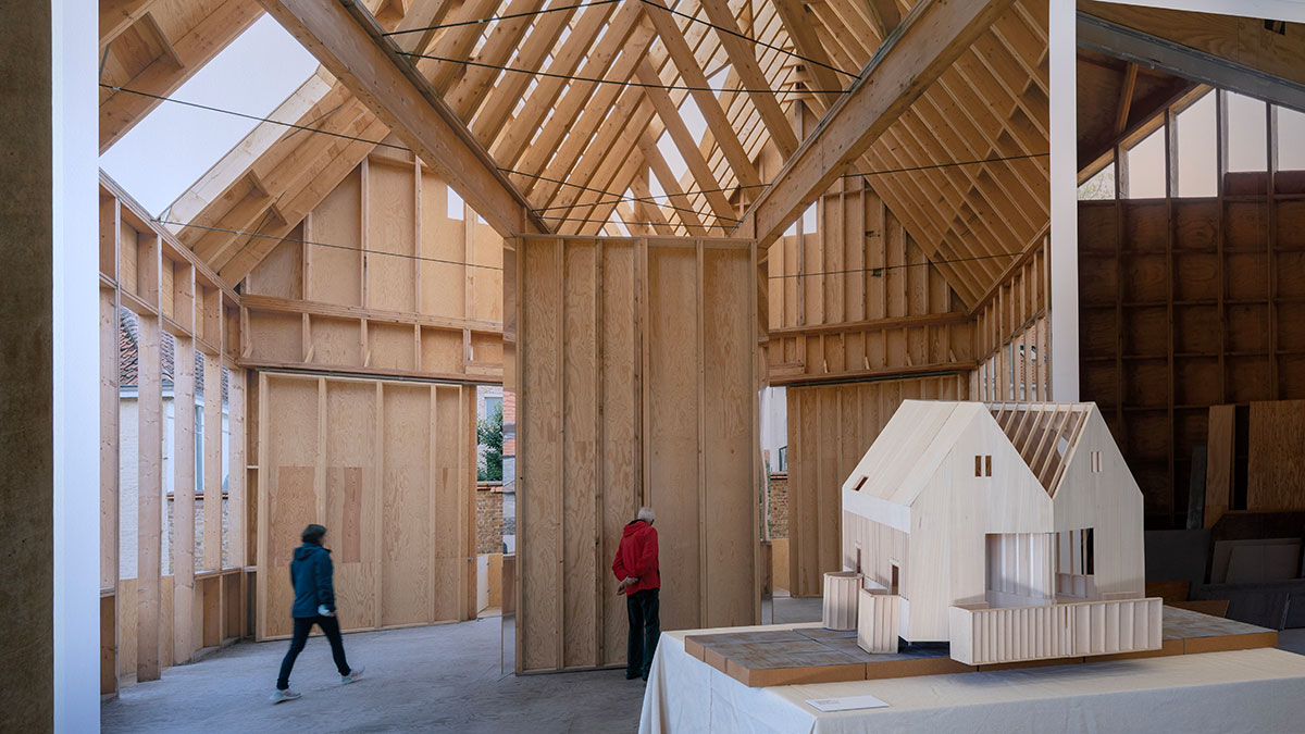 A wooden architectural model on a pedestal in front of a large wall mural image of people standing inside a wooden building.