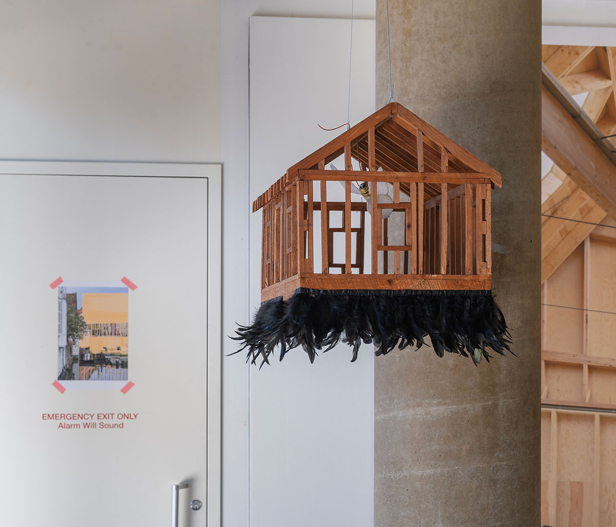 A wooden architectural model with black feathers attached to the bottom suspended from the ceiling.