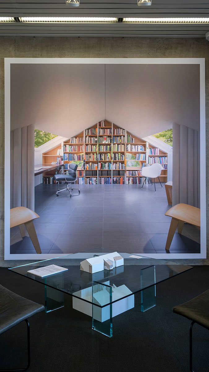 An architectural model on a glass table in front of a wall mural image of an interior room with seating and bookshelves.