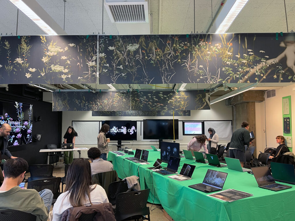 A final review for a course in a classroom filled with students and instructors. Rows of laptops sit on a table covered in a green tablecloth. On the ceiling are digital renderings of plants.