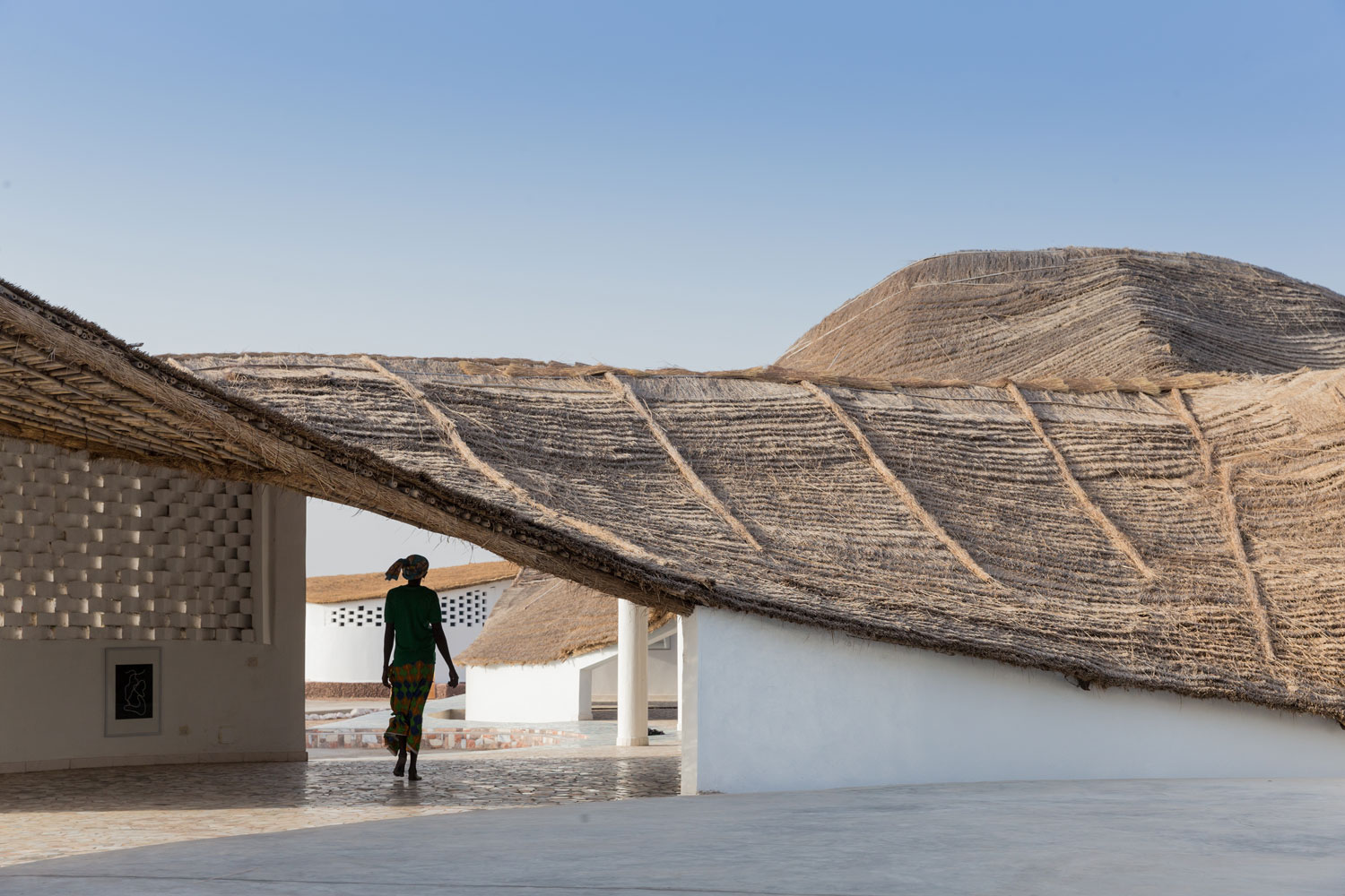A woman walks below a low porch-like strucuture of organic material beside a white building.