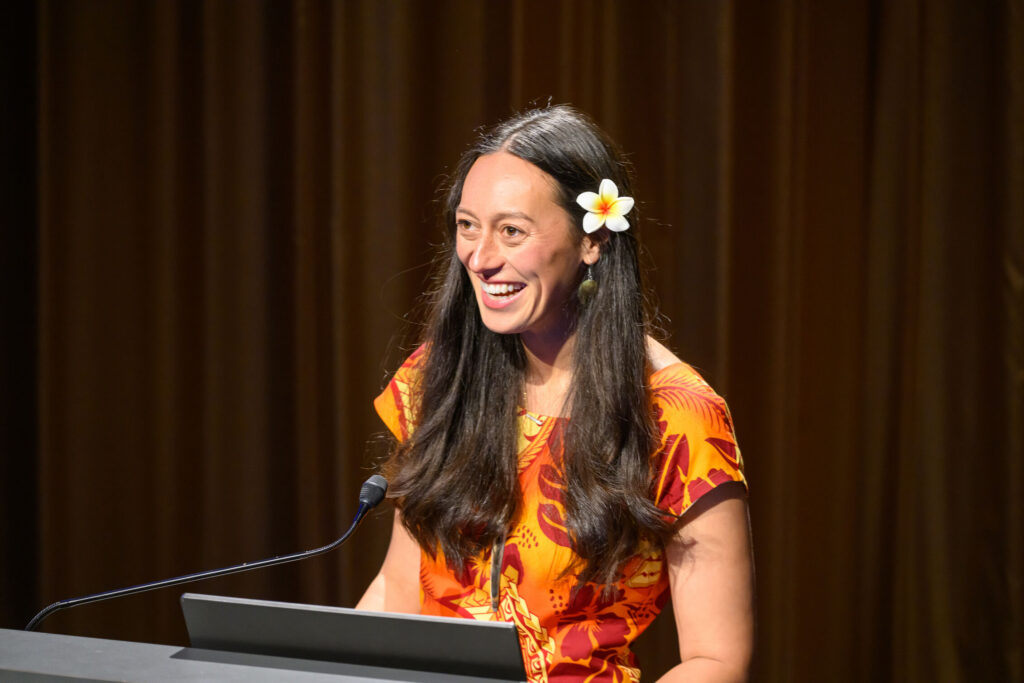 Elyjana Roach stands behind a podium in an orange dress and flower in her hair smiling