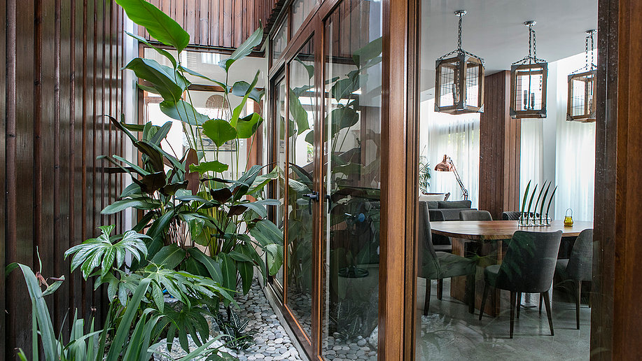 Indoor courtyard with tall green plants and white pebbles enclosed by glass walls, opening into a modern dining room with a wooden table, dark upholstered chairs, and hanging lantern-style light fixtures.