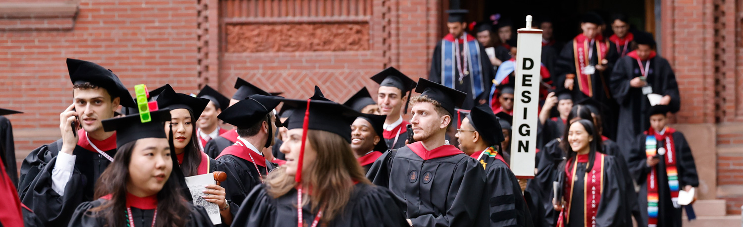 Many people in In front of a brick building, graduation gowns and caps with a vertical sign that says "Design"