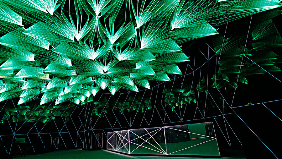 Dark futuristic interior with a ceiling of glowing green geometric light installations and mirrored walls reflecting the angular patterns.