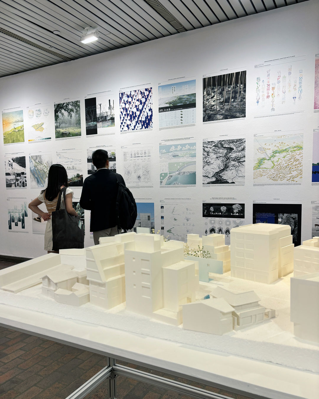 Two people look at student work on a wall at the GSD. An architecture model is in the foreground.