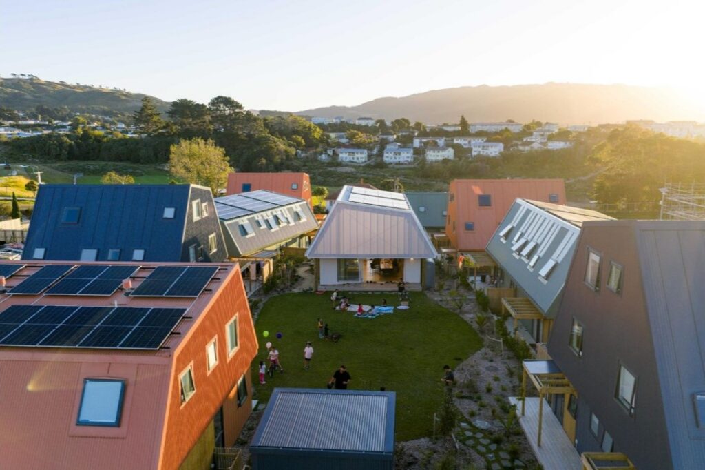 Aerial view of a modern cohousing community at sunset, with colorful, angular townhouses surrounding a shared grassy courtyard where a few small groups of people and children play and relax on picnic blankets; several roofs are covered with solar panels, and green hills and a residential neighborhood stretch into the background.
