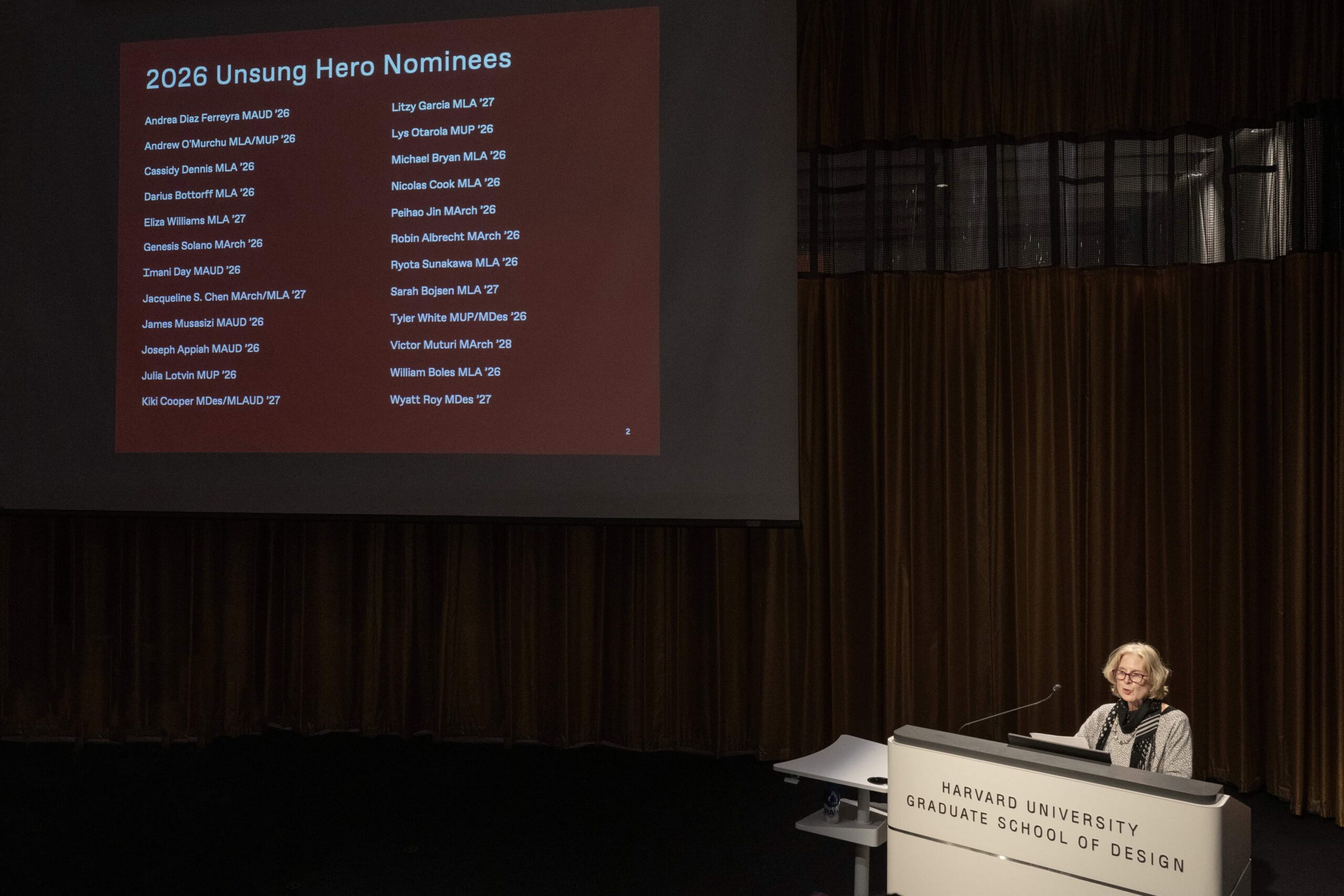 A woman stands behind a podium. Behind them, a projector shows a slide that reads "2026 Unsung Hero Nominees"