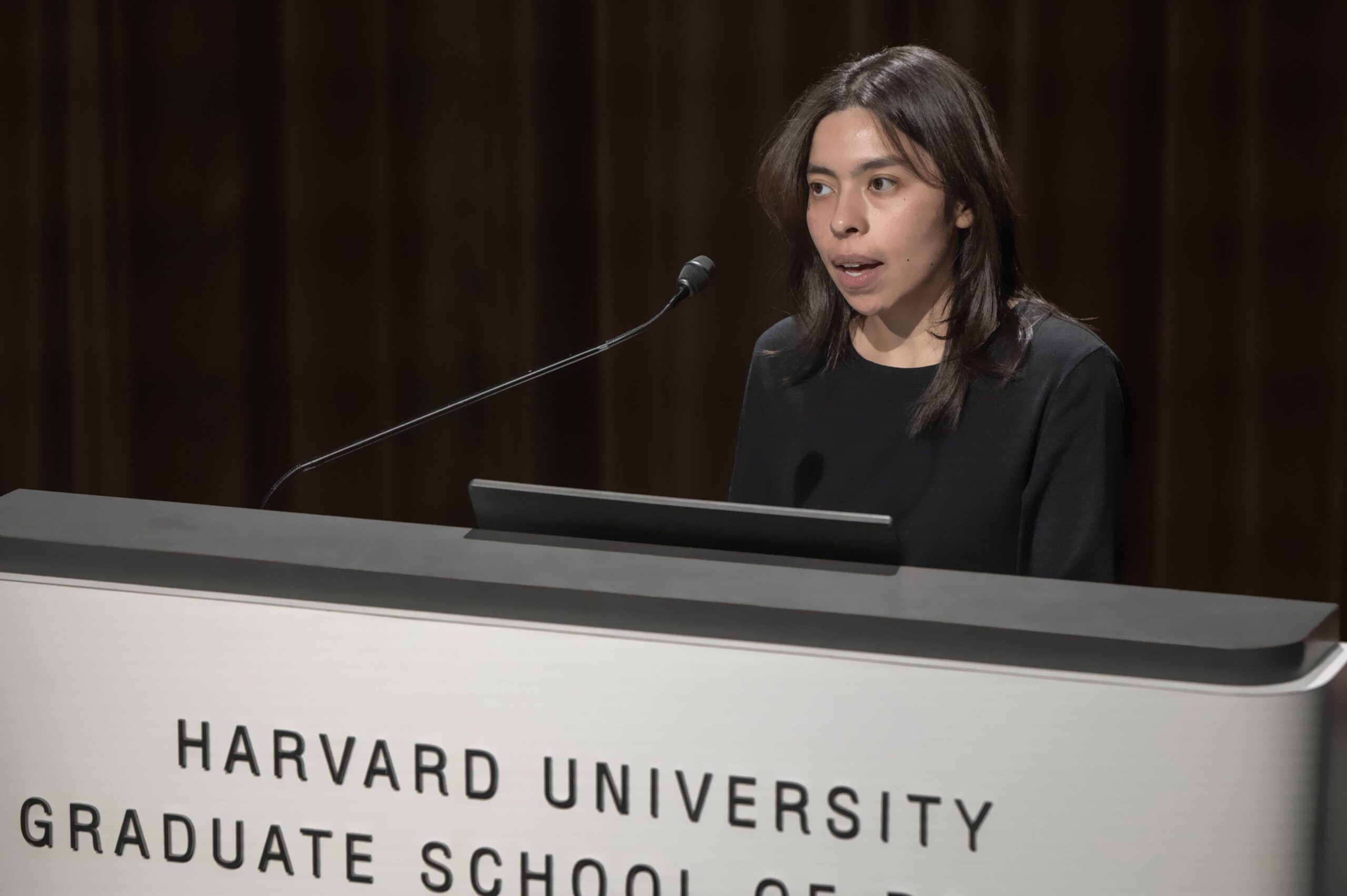 A person with long black hair stands behind a podium that reads "Harvard University"