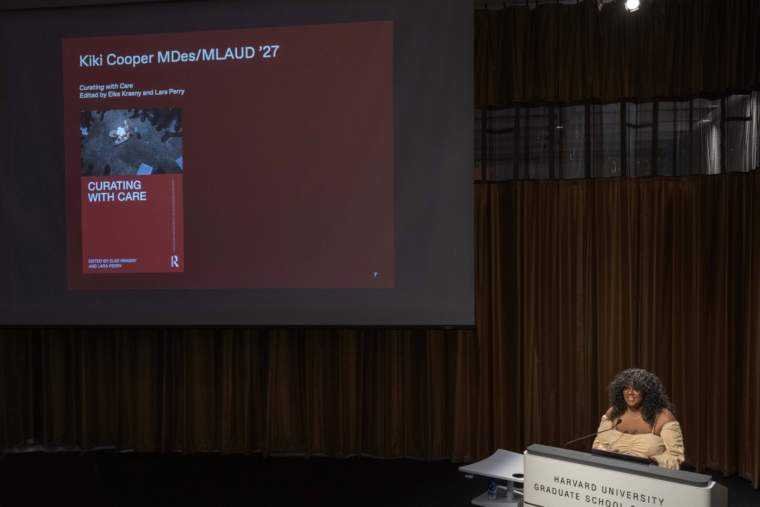 A person with curly hair stands behind a podium with a spotlight on them. Behind them, a projector shows a slide that reads "Curating with Care."