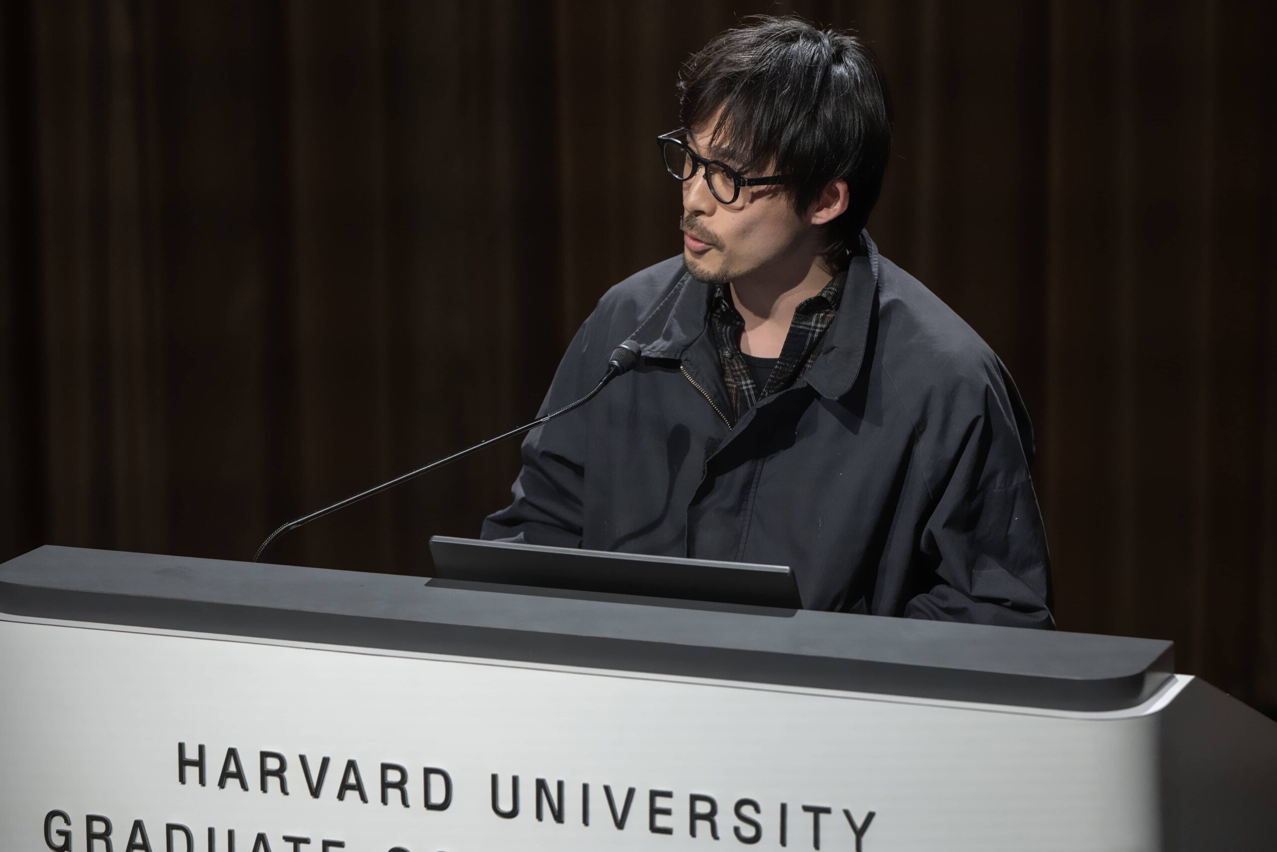 A man with black hair and glasses stands behind a podium.