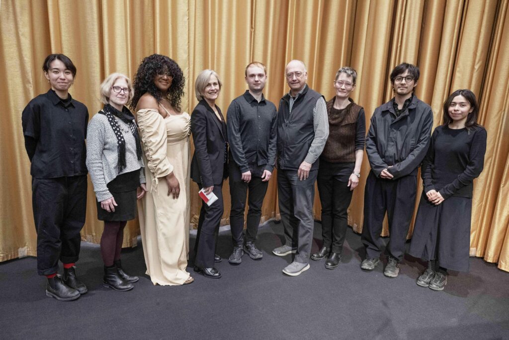 Nine people stand in a line posing for a picture in front of a gold curtain