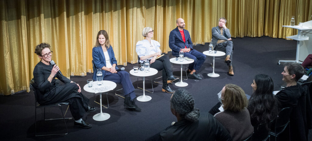 Five speakers sit on a stage in front of a gold curtain, addressing an audience.