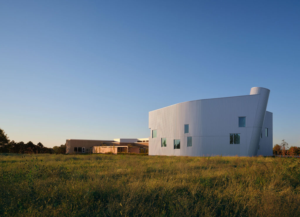 Contemporary white building with curved metal façade rising from a grassy field under a clear sky.