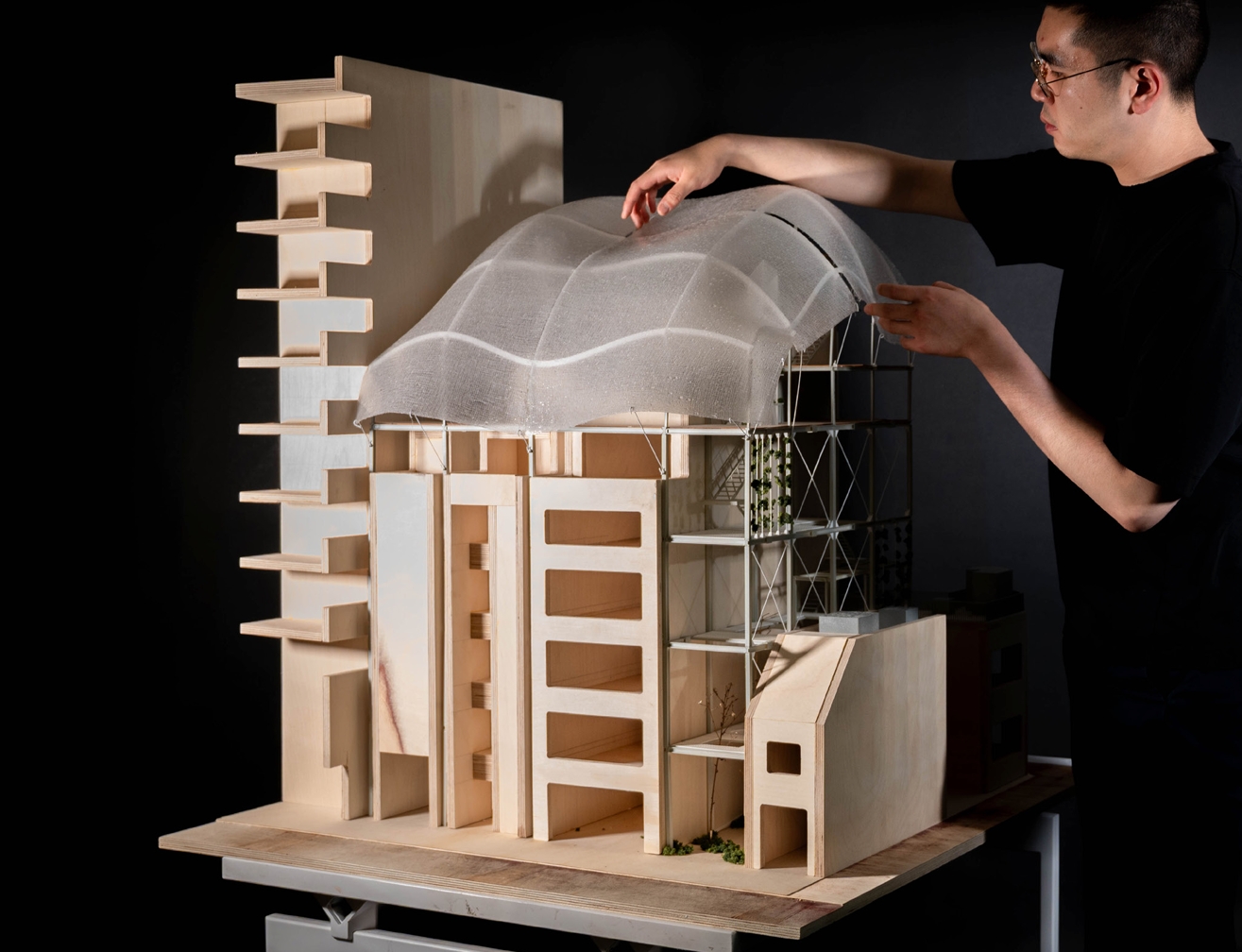 Student adding finishing touches to mixed media architectural model against black background.