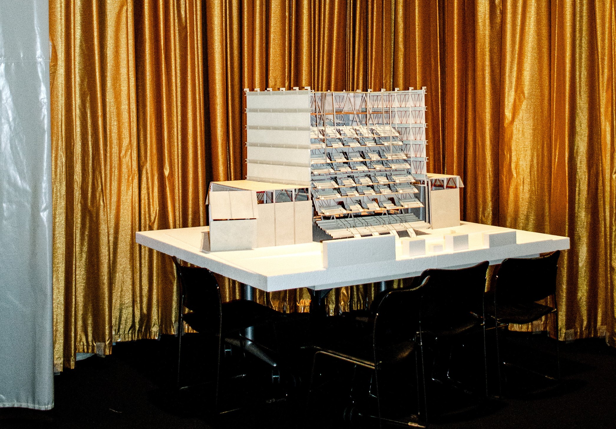 Architectural model of box-like stadium structure, posed in front of gold curtain.