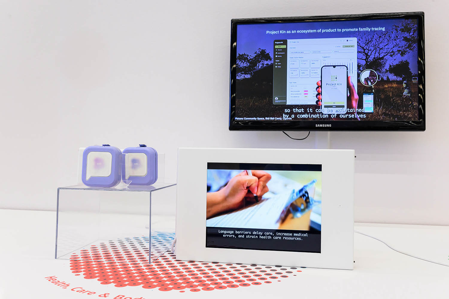 A display showing two small devices on an acrylic pedestal and a small screen showing how the device is used. Behind is a larger monitor showing another project.