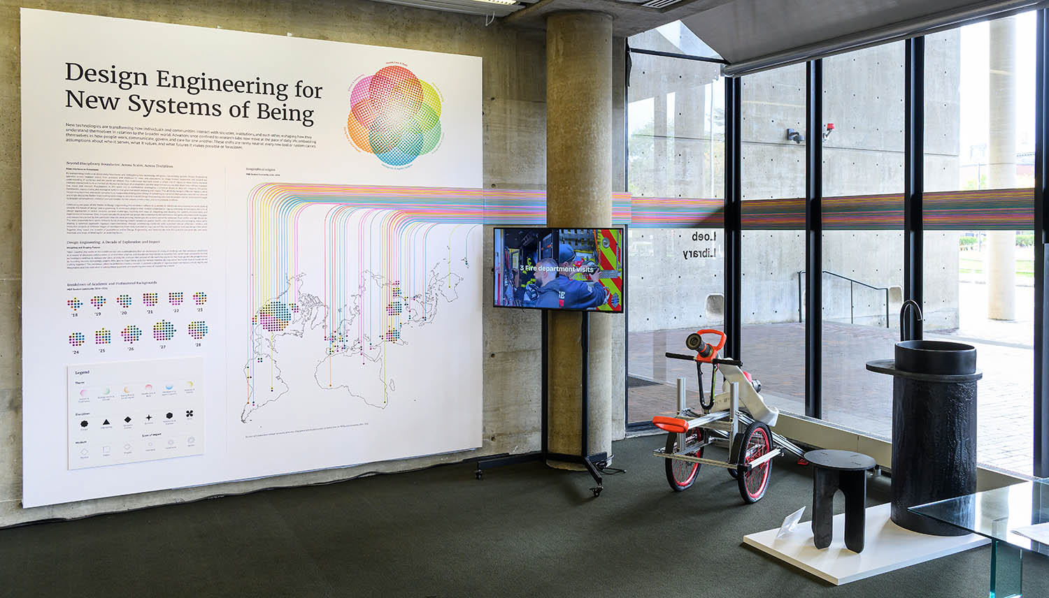 Introduction wall with a monitor on a stand against a column. There is a device on bicycle wheels holding a fire-hose and black table and black sink.