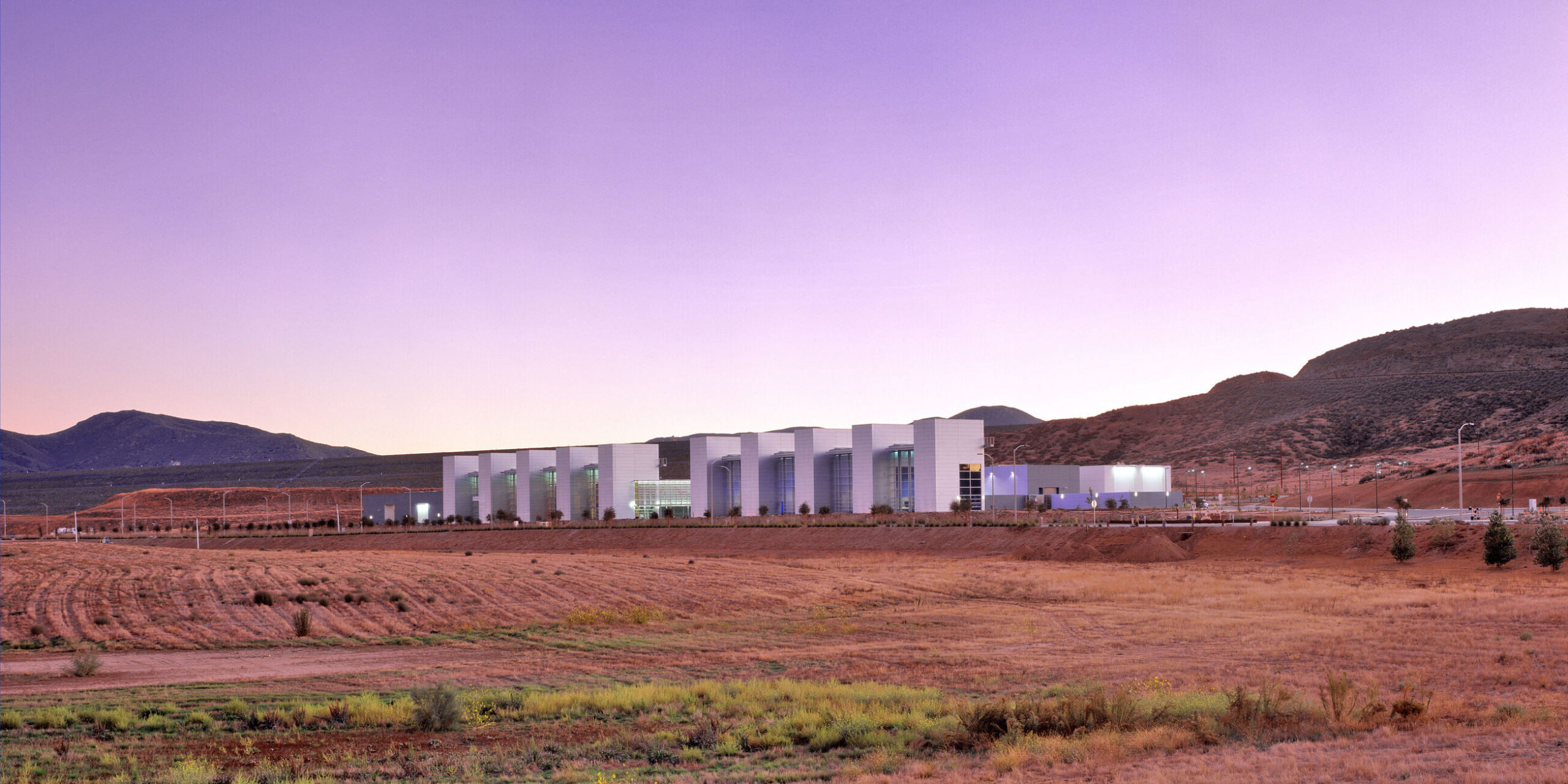 Wide panorama of a long, low, white building in a dry valley, framed by scrubby fields in the foreground and low mountains under a pink‑purple twilight sky.