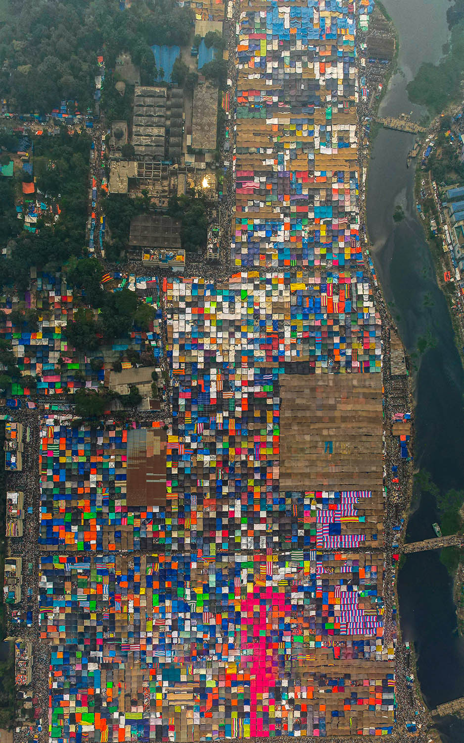 A top-down view of a colorful grid of square tents betweem a city and a river.