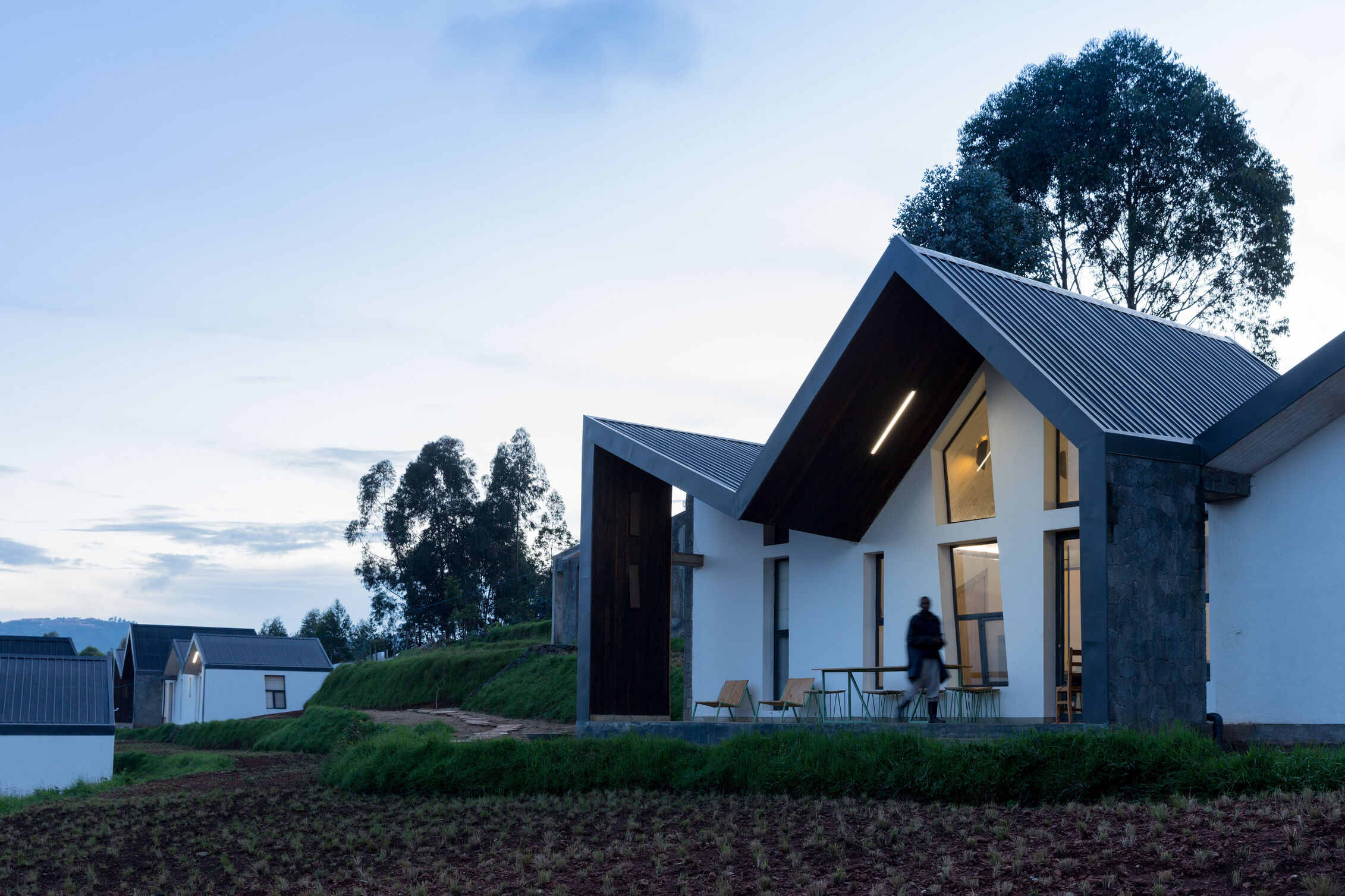 Evening view of a modern white house with steep angular roofs and large windows, set on a green hillside with other small houses and tall trees in the background.