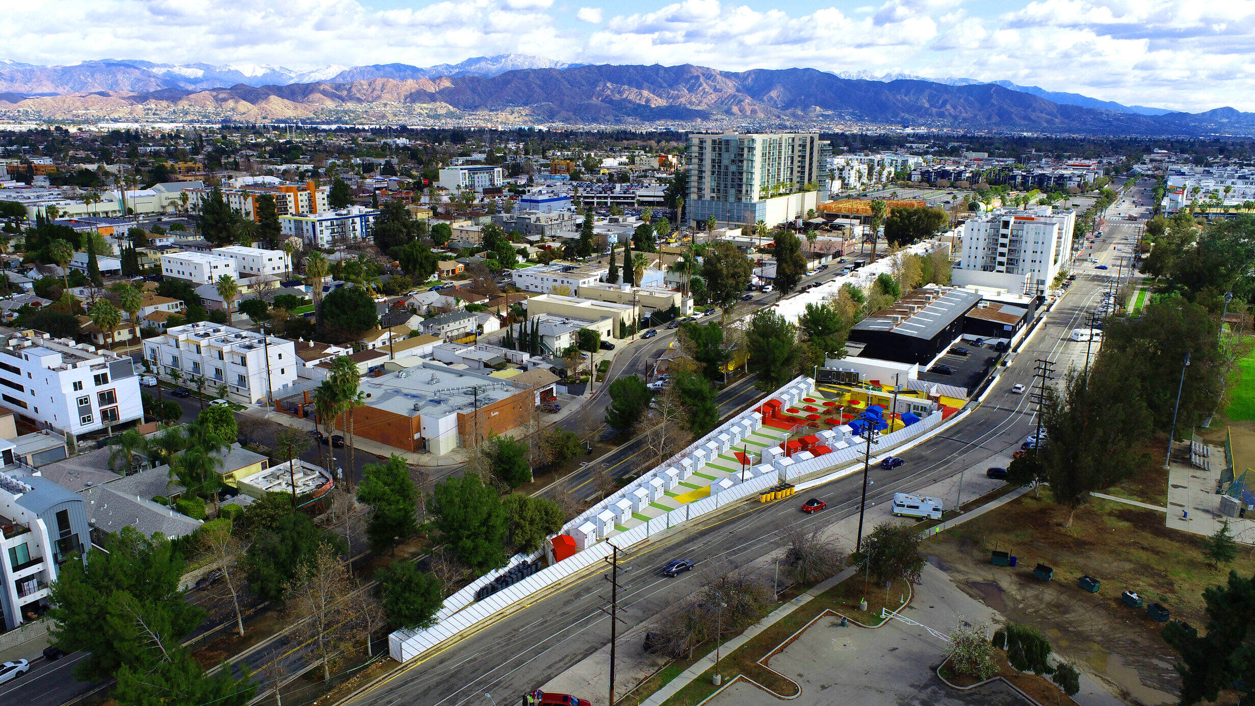 Aerial view of Los Angeles featuring low‑rise buildings and tree‑lined streets, snow‑capped mountains in the distance, and a long row of small colorful tiny homes along a curved roadside.