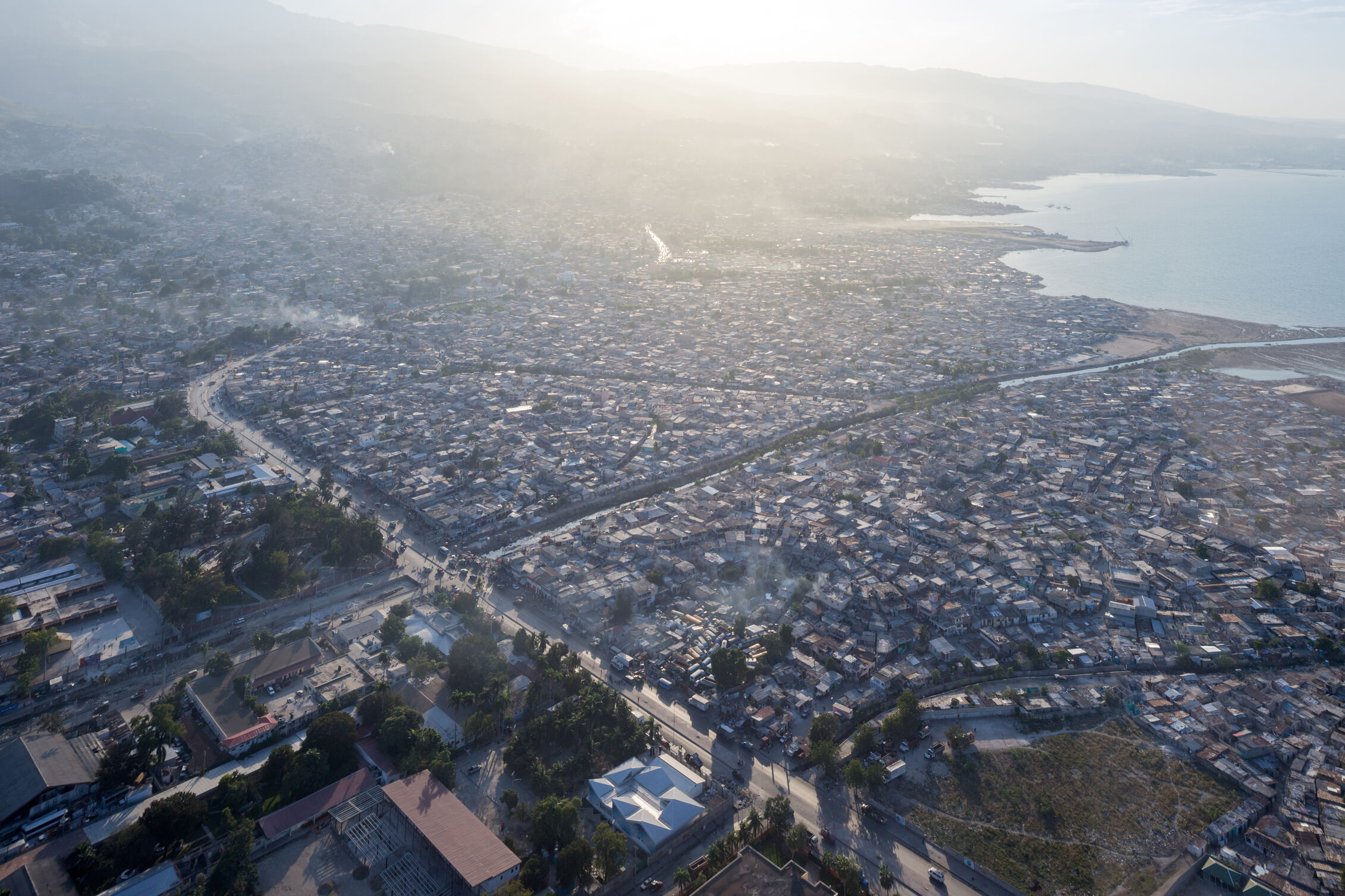 Aerial view of a dense coastal city at sunrise or sunset, hazy light washing over tightly packed low‑rise buildings stretching from the shoreline to distant hills.