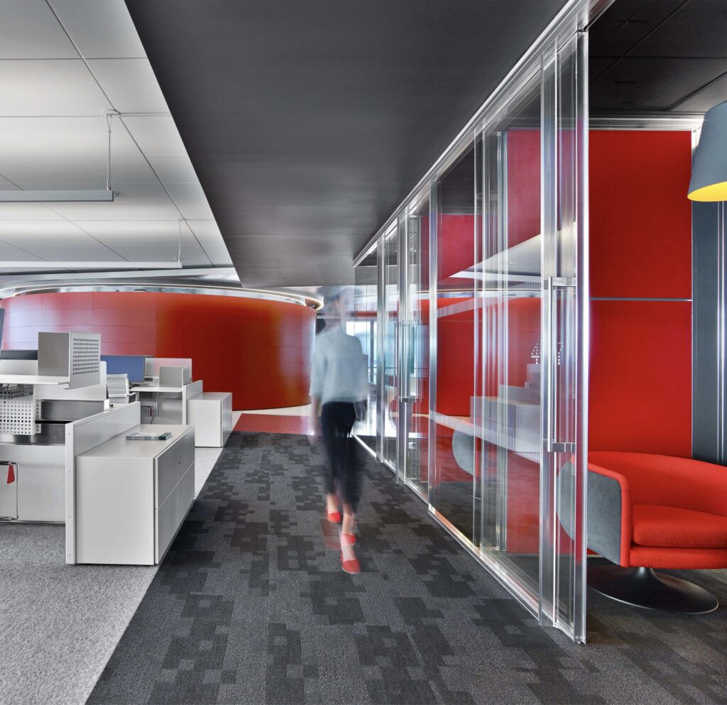 Contemporary office interior with gray and patterned carpet, white workstations on the left, glass partitions and bright red curved walls and seating on the right, and a blurred person walking down the corridor.