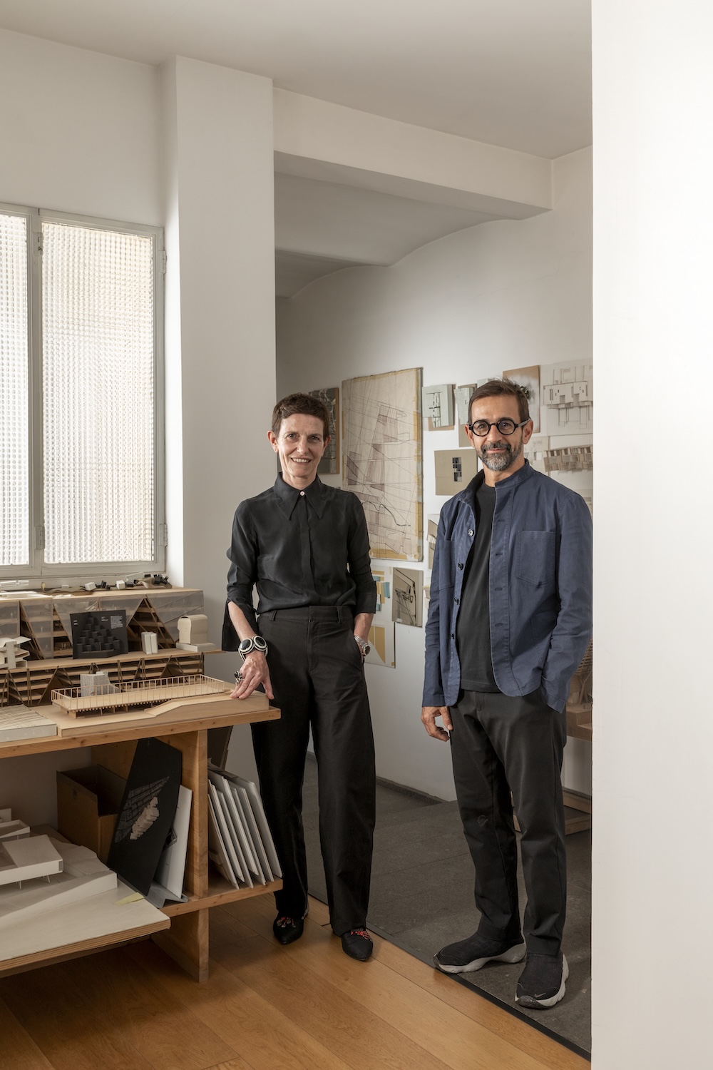 Maria Claudia Clemente and Francesco Isidori stand smiling in an architecture studio, surrounded by shelves of scale models, sketches, and drawings pinned to the wall, with a wooden floor and large window in the background.